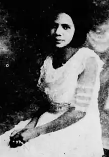 A young African-American woman, seated with her hands clasped in her lap. She is wearing a white lacy dress.