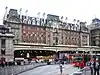 A grey building with three rectangular, white signs reading "London Victoria Station" in black letters all under a clear, white sky