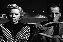 Black-and-white image of a man and woman, both with downcast expressions, sitting side by side in the front seat of a convertible. The man, on the right, grips the steering wheel. He wears a jacket and a pullover shirt. The woman wears a checkered outfit. Behind them, in the night, the road is empty, with a two widely separated lights way off in the distance.