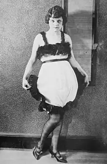 A young white woman with dark bobbed hair, standing, wearing a short light -colored sleeveless dress with dark fringe and a bubble-style skirt