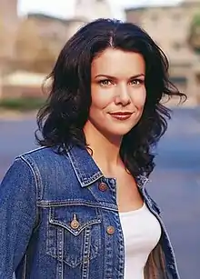 A young brunette woman smiling towards the camera, wearing a denim jacket over a white tank top.