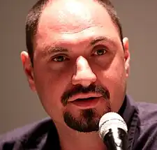 A man plays a guitar, while the symbol of his band is viewable behind him.