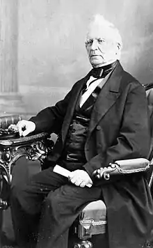 Phot of fair-skinned man with white hair, tuft in front, wearing mid-Victorian business suit, sitting at a table