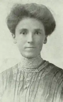 A white woman with dark hair in a bouffant updo, wearing a high-collared blouse or dress with lace trim and pintucks