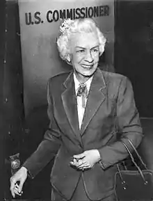 Photograph of a white-haired slender woman in a business suit carrying a purse on her left arm.