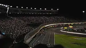 A photo of NASCAR cars racing at the [[2008 Bank of American 500]] during night time.