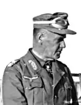 Crüwell is seen in profile. He wears a military uniform, along with a cap for desert warfare. His Iron Cross displayed at the front of his shirt collar.