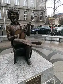 Statue of Luke Kelly sitting on a chair and playing an instrument and (presumably singing). The statue is placed on a cement platform. The platform is on a pavement with benches to sit on. In the background there is a big brown building, with cars and bicycles parked in front, along with tree (without leaves). There are other smaller buildings on top right corner away from focus.