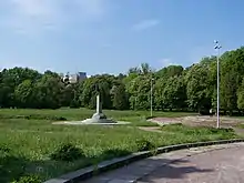 A green park with a statue in the middle of the picture. The statue has three small steps up to its base, which is a half football. There is then a column leading up to a bronze-brown falcon statue. It is difficult to see the details but the falcon is on top of a football as well.