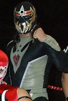  A masked professional wrestler in the ring, wearing a black mask and black bodysuit, both with gold markings