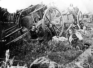 A black-and-white photograph showing a group of wooden carts sitting in a field with people resting in the grass and on the carts.