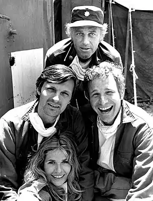 A black-and-white photograph of three men and one woman on a bright day all looking at the viewer and wearing dark, collared jackets