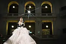 A person in a voluminous white dress and black gloves with the arches of the La Trobe Reading Room in the background