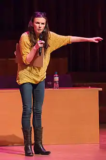 A white lady with long hair, is wearing yellow blouse, blue pants, and black boots. She is holding a microphone with her right hand and talking on it. Her left hand is stretched out. There is a wooden table on the background with a bottle of water on it