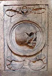 Close-up of a rectangular-shaped carving in stone. In the centre of the rectangle is a circle representing a mirror, and within the circle is a grinning skull. The circle is framed by ram's horns.