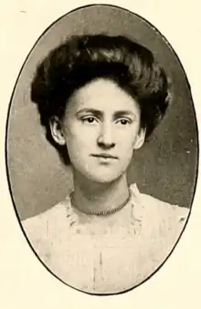 A young white woman with dark hair in a bouffant style, wearing a white dress with a square, lace-trimmed neckline, and a choker-style strand of beads