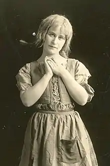 A young white woman with blonde hair cropped short, wearing a folk-inspired costume with laced closure and an apron with a pocket. Her hands are clasped against her chest.