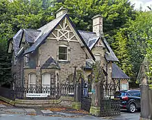 An intricate stone structure, two stories high, with gabled roofs and half-timbering in the gable apexes. To its right, and closer to the camera, is a wrought iron fence and gates with intricate, pyramid-topped stone gateposts. A sign on the fence in front of the building says "crematorium".