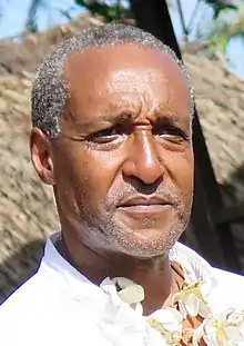 Half portrait of a man at a 3/4 angle in direct sunlight, wearing a white shirt with a necklace of white flowers. In the background is a wooden porch and turquoise house in front of a more distant grass roof.