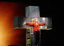 Madonna standing on a blue crystal cross, wearing a red shirt and dark brown pants. Her hands are spread apart along the cross's breadth as if she has been crucified. Behind the cross, a backdrop is centrally illuminated