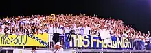 A crowd wearing white, blue and yellow cheers in bleachers