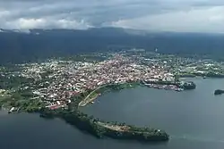 In the distance are cloud covered mountains, while in the front of the picture are houses leading to the harbour which has two piers leading out to sea