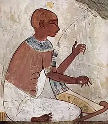 A brown-skinned man in white-linen garb, seated and playing a stringed harp with both hands