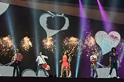 A woman performing on a stage in front of white hearts alongside five men.