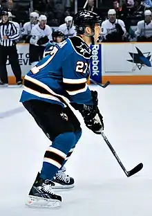 An ice hockey player, seen from the side, in a ready position. He is slightly crouched while standing on his skates and holds his stick in both hands. He wears a teal jersey with black trim, as well a black helmet.