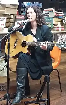 A photograph of Manon Steffan Ros sat in a bookshop, speaking to an audience with a guitar placed on her lap and a microphone in a stand at her side.