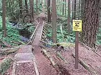 A trail and boardwalk through trees and brush with a caution sign next to the trail