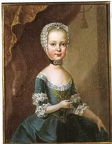 A young, blonde girl in a greenish-blue dress and a white cap. Her dress is embellished with white lace, greenish-blue bows and golden embroidery.