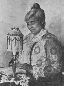 An older African-American woman with grey hair, seated at a table, hands and eyes on an open book; there is a small ornate lamp nearby, and lace on the tablecloth. She is wearing a lace or embroidered dress.