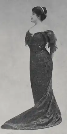 A white woman, standing, with dark hair in a bouffant updo, wearing a dark gown with bare shoulders, lace sleeves, and a long train.