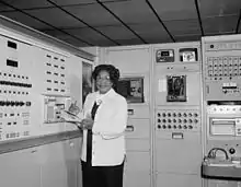 Black and white photograph of Mary Jackson standing in front of large instruments, holding a clipboard and pencil