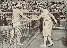 Lenglen and Wills shaking hands at the net while an official at the side holds their right hand up in a "stop" gesture