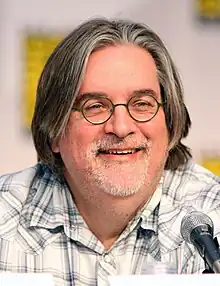 A smiling man in glasses and a plaid shirt sits in front of a microphone.