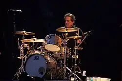 Middle-aged man with slightly graying dark hair and glasses wearing black shirt sits behind a silver-colored, gold-lit drum kit with his left hand and stick about to hit a cymbal and his eyes fixed straight ahead.