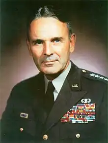 Middle-aged man with greying dark hair parted slightly off-centre. He wears a green dress uniform, with suit and tie, is clean-shaven, and has four stars on his shoulder to indicate his rank.