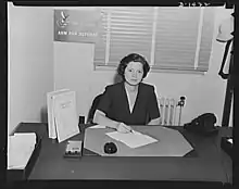 Photograph of a woman with short hair wearing a dark-colored v-necked dress, sitting at a desk in a government office