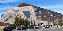 An angular reddish-gray building stands before a brown building on a clear day.