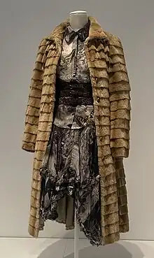 A light brown fur coat worn over a blouse and skirt in a fabric printed with birds, moths, and skulls