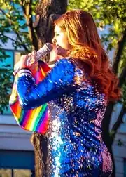 A woman with brown hair holding a microphone and a rainbow flag