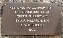 Colour photograph of the brass plaque with the commemoration written in upper case on white lettering. The commemoration reads as follows: "Restored to commemorate the Silver Jubilee of Queen Elizabeth II by A. R. Millard B.E.M. & volunteers 1977."