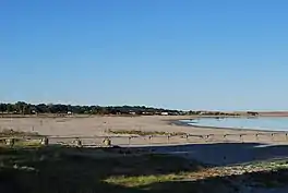 The picture shows Lake Albert from its shore. In the foreground is a sandy shore and a pipeline leading towards the lake. The lake is in the right of the photo. In the background there are trees on the left and brown hills on the right.