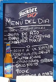 A board advertising Buckler 0,0° beer. It lists: MENÚ DEL DIA. - REV. CHAMPIÑONES - SOPA DE AJO - SOPA DE PICADILLO - ENSALADA MIXTA -○- - LOMO A LA BRASA - COSTILLA EN SALSA - FILETES RUSOS -CALAMARES SALSA - GALLO FRITO. 9'00 €. BEBIDA APARTE