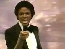 A adult African American male with short black hair. He is pointing forward with his right hand and is making a facial expression and he is wearing a black bow tie with a black and white tuxedo. Behind him, there is a pink background.