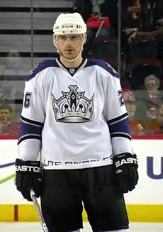 An ice hockey player standing facing towards the camera. He is wearing a white helmet, and a white, black, and purple uniform with a large crown on his chest.