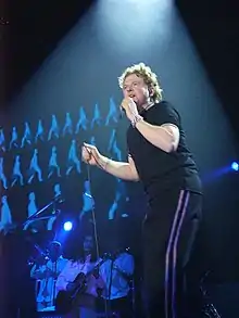 A man wearing a black T-shirt and black trousers singing into a microphone.