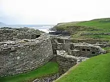 A semi-circular stone wall at left hints at the existence of a large and ancient building and to the right are the ruins of various other stone structures. In the background a low cliff divides a body of water from grassy fields.